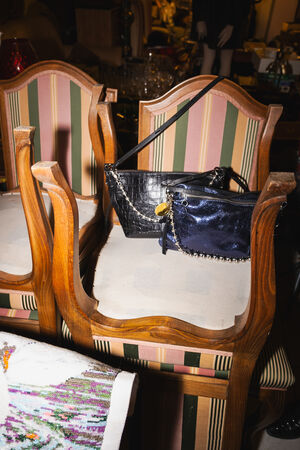 Zwei Holzstühle mit gestreifter Polsterung sind übereinander gestapelt und präsentieren zwei INA KENT Handtaschen - eine schwarze und eine dunkelblaue - auf der Sitzfläche. Ein gemusterter Stoff ist über die Armlehne drapiert und verleiht ihr einen Hauch von Charme.