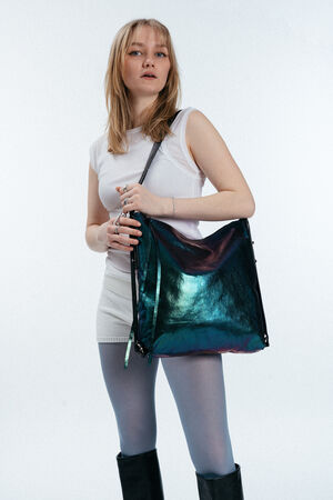 Frau in weißem Oberteil und Rock, blauer Strumpfhose und schwarzen Stiefeln, die eine schillernde große INA KENT-Umhängetasche vor einem schlichten hellen Hintergrund hält.