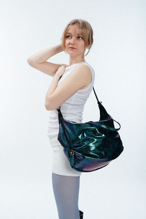 Eine Frau in einem weißen ärmellosen Oberteil und Rock steht mit einer großen schillernden INA KENT-Umhängetasche vor einem schlichten weißen Hintergrund und schaut zur Seite.