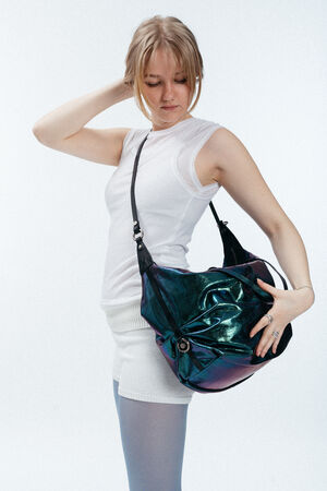 Eine Frau in einem ärmellosen weißen Oberteil und kurzen Hosen posiert mit einer großen, glänzenden, schillernden INA KENT-Umhängetasche vor einem schlichten, hellen Hintergrund.