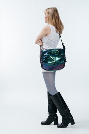 Eine Person, die ein weißes Oberteil, eine hellblaue Strumpfhose und schwarze Stiefel trägt und eine glänzende, schillernde INA KENT-Tasche bei sich hat, steht vor einem schlichten weißen Hintergrund.