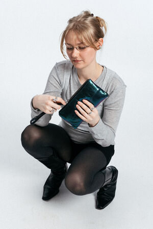 Eine Frau mit Brille und grauem Oberteil geht in die Hocke, schaut in ihre glänzende INA KENT-Tasche und hält eine Schere in der rechten Hand.