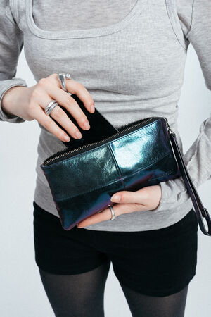 Eine Person, die ein graues langärmeliges Hemd und schwarze Shorts trägt, legt ein schwarzes Smartphone in eine glänzende, schwarze INA KENT-Clutch-Tasche mit Reißverschluss.