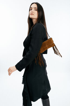 Woman with long dark hair wearing a black coat and holding an INA KENT brown fringed shoulder bag, walking against a plain white background.