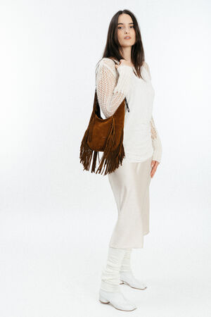 Woman standing against a white background, wearing a cream sweater, light skirt, white boots, and carrying an INA KENT brown fringed shoulder bag.