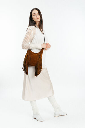 A woman wearing a white long-sleeve top, cream-colored skirt, white boots, and leg warmers poses with an INA KENT brown fringed shoulder bag against a plain white background.