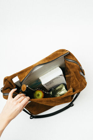 A hand opens a brown suede INA KENT bag containing books, an apple, and a metallic pouch, viewed from above on a white background.
