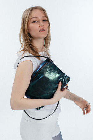 A woman with blonde hair wearing a white outfit holds a large, shiny dark teal INA KENT bag under her arm against a plain light background.