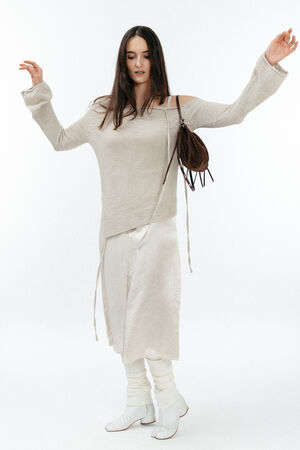 Woman in an off-the-shoulder beige sweater, white skirt, leg warmers, and white shoes holds an INA KENT brown bag, standing against a plain white background.