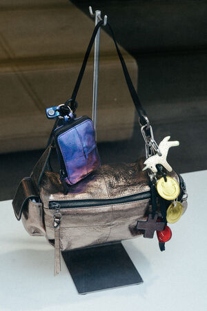 Eine INA KENT Metallic-Crossbody-Tasche mit einem kleinen schillernden Beutel und verschiedenen Schlüsselanhängern, darunter ein Vogel, Münzen und ein Kreuz, die an ihrem Riemen befestigt sind.