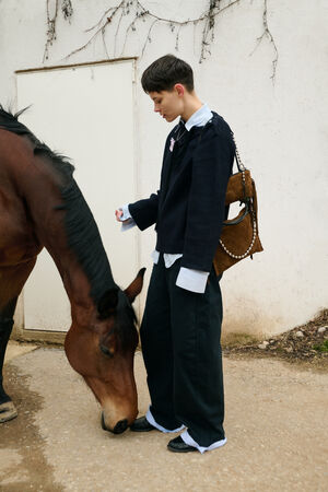 Eine Person in lockerer, mehrschichtiger Kleidung steht neben einem braunen Pferd mit gesenktem Kopf, das vor einer weißen Wand den Boden neben seinen Füßen beschnüffelt, und trägt eine INA KENT-Tasche.