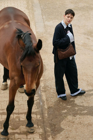A person in loose-fitting clothes stands on a paved area holding an INA KENT bag, next to a brown horse walking away.