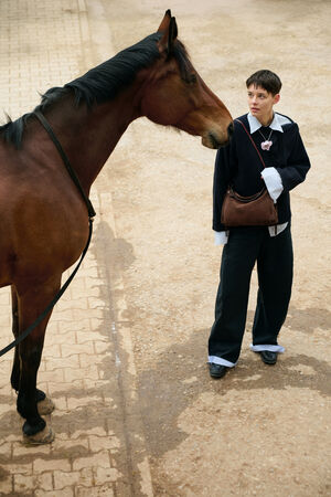 A person wearing loose black pants and a dark sweater, carrying an INA KENT bag, stands on a paved path, looking at a brown horse standing nearby.
