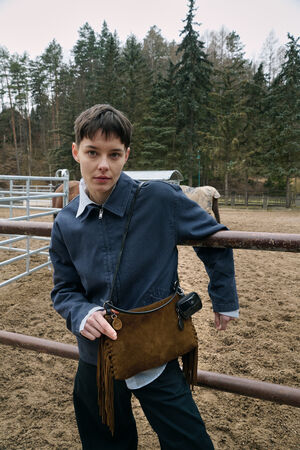 Person with short hair leans against a metal fence in an outdoor enclosure, wearing a blue jacket and holding a distinctive INA KENT brown fringed bag; trees and fencing visible in the background.