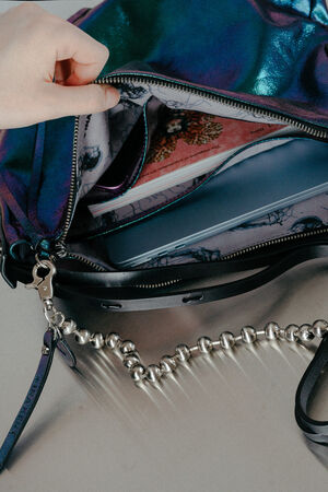 A hand opens an INA KENT zippered, iridescent handbag revealing a laptop and a pink notebook inside. A metallic beaded strap is attached to the bag.