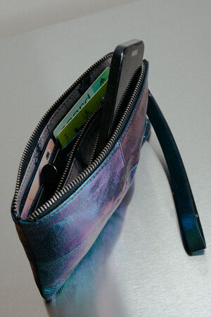 Eine INA KENT Clutch mit Reißverschluss und Handschlaufe, die ein Smartphone, Bargeld, Karten und einen Lippenstift enthält, ruht elegant auf einer Metalloberfläche.