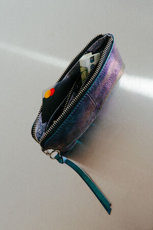 Eine kleine, schillernde INA KENT-Brieftasche mit Reißverschluss ist teilweise geöffnet und offenbart eine Mastercard-Kreditkarte und Bargeld, die auf einer hellen Oberfläche liegen.