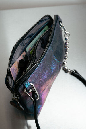 Eine kleine schillernde INA KENT Geldbörse mit Reißverschluss ist geöffnet und offenbart ein paar Karten, Bargeld und einen Lippenbalsam im Inneren. Ein Kettenriemen ist an der Geldbörse befestigt.