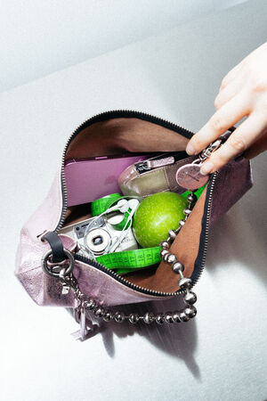 A hand holds open a pink INA KENT purse containing a green apple, a camera, a tape measure, a phone, and a coin purse on a silver surface.