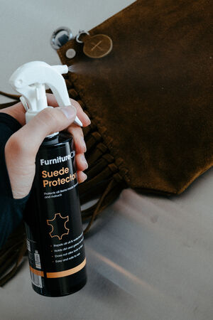 Eine Person hält eine Dose Furniture Clinic Suede Protector Spray neben einer braunen INA KENT Wildledertasche auf einem Tisch.