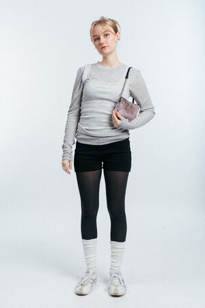 Person standing against a light background, wearing a grey long-sleeve shirt, black shorts, black tights, white socks, sneakers, and carrying a small purple INA KENT shoulder bag.