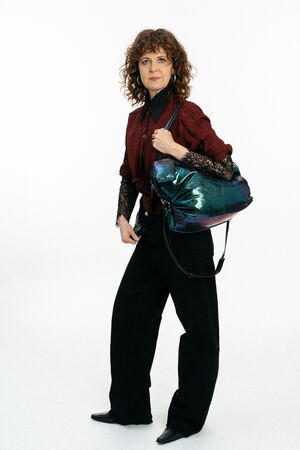 Eine Frau mit lockigem Haar, burgunderrotem Hemd und schwarzer Hose steht mit einer großen, schillernden INA KENT-Tasche über der Schulter vor einem weißen Hintergrund.