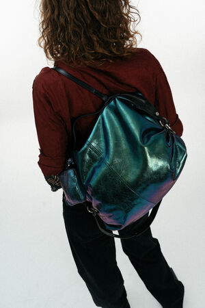 Eine Person mit krausem Haar, kastanienbraunem Hemd und schwarzer Hose trägt einen INA KENT-Rucksack in metallisch glänzendem Teal auf dem Rücken vor einem schlichten weißen Hintergrund.