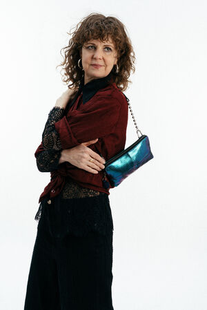 Frau mit lockigem Haar in roter Bluse, schwarzem Spitzentop und schwarzer Hose trägt eine schillernde Handtasche von INA KENT auf ihrer Schulter vor einem schlichten weißen Hintergrund.