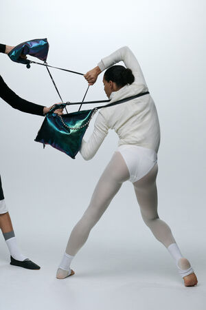 Zwei Personen ziehen an einer großen, schillernden INA KENT-Handtasche und scheinen sich darum zu streiten; beide tragen helle, figurbetonte Kleidung vor einem schlichten Hintergrund.