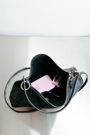 Eine schwarze INA KENT-Handtasche mit offenem Reißverschluss offenbart ein rosafarbenes Gerät, eine Sonnenbrille und verhedderte Kopfhörer, die alle auf einer hellen Fläche liegen.