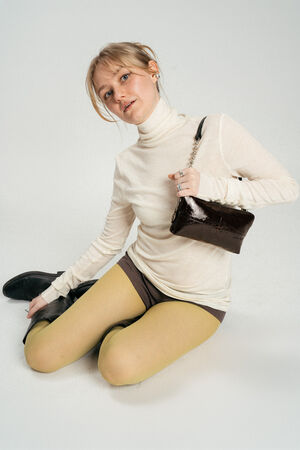 Person sitting on the floor wearing a cream turtleneck, yellow tights, shorts, black boots, and holding a chic INA KENT black handbag.