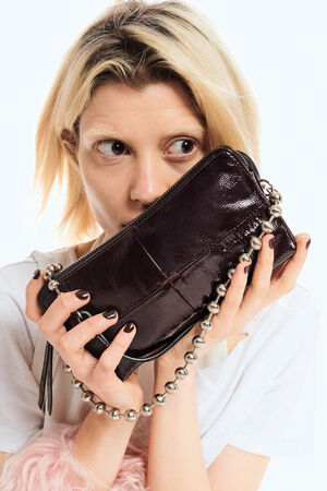 A person with blonde hair holds an INA KENT small, dark patent leather handbag with a beaded strap near their face.