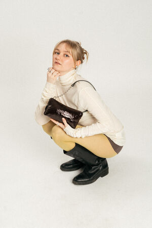 A person in a cream turtleneck, yellow tights, brown skirt, and black boots squats while holding a small dark INA KENT handbag against a plain white background.