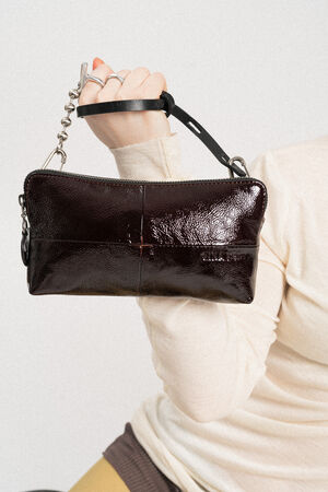 A person holds up a small, black INA KENT patent leather handbag with a chain strap against a plain background.