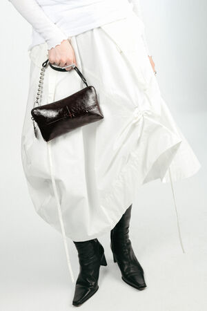 Person wearing a white skirt and black pointed boots, holding a chic INA KENT black handbag with a chain strap, against a plain light background.