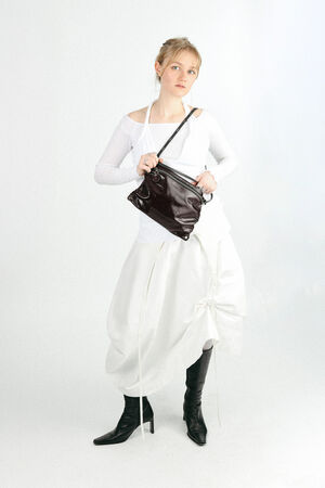 A person in a white long-sleeve top and white skirt stands holding an INA KENT black shoulder bag, wearing black heeled boots against a plain white background.