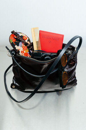 A black INA KENT handbag on a table contains a floral eye mask, a comb, a red passport holder, a camera, and sunglasses.