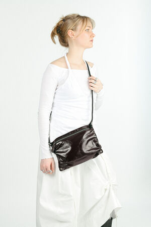 A person wearing a white long-sleeve top and skirt stands against a white background, holding a shiny black INA KENT crossbody bag.