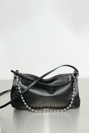 Schwarze Lederhandtasche von INA KENT mit Reißverschluss, passendem Schulterriemen und einer dekorativen Kette mit runden Silberperlen, die auf einer glatten Oberfläche ausgestellt ist.
