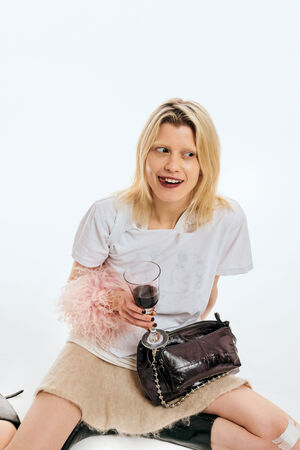 A woman with blonde hair sits on the floor holding a glass of red wine, wearing a white t-shirt, beige skirt, and boots, with an INA KENT black purse and a pink feather accessory.