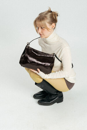 A person in a white turtleneck and yellow pants squats while holding a large, shiny black INA KENT handbag.