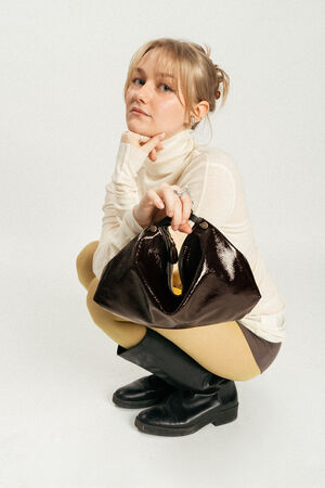 A woman in a white turtleneck, yellow tights, and black boots squats while holding a shiny black INA KENT handbag against a plain background.