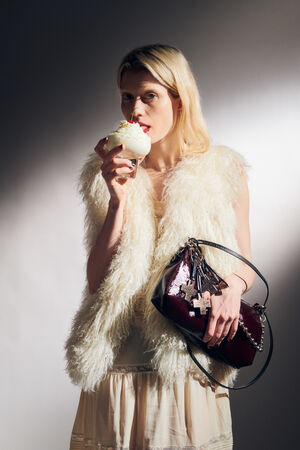 A woman wearing a white faux fur vest holds a dessert with whipped cream and an INA KENT dark purple handbag, standing against a plain background.