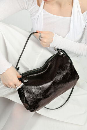 Person in white clothing holds a shiny black INA KENT leather handbag, partially unzipped, while seated against a white background.