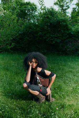 A person with curly hair crouches on green grass, resting their face on one hand, in front of dense green foliage, with an INA KENT bag by their side.