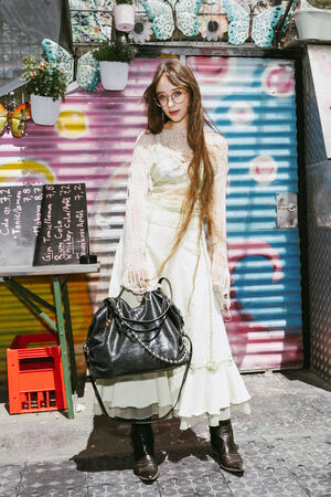 A person with long hair, glasses, and a lace top over a pale dress stands holding a large black INA KENT bag in front of a colorful mural and hanging potted plants.