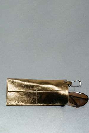 A metallic gold INA KENT eyeglass case with a carabiner clip, partially open, revealing a pair of brown sunglasses inside.
