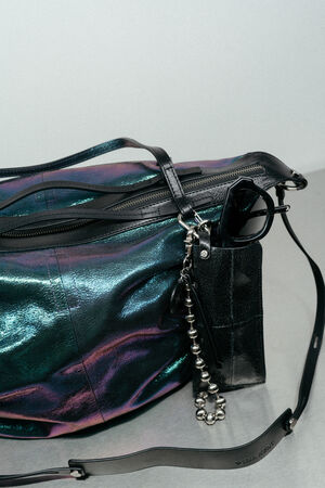 A holographic black INA KENT shoulder bag with a zipper, black sunglasses, a small black pouch, and a metallic beaded keychain on a gray surface.