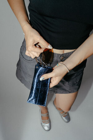 Person holding a pair of sunglasses and a shiny blue INA KENT case, wearing a black top, gray skirt, and silver shoes, photographed from above.