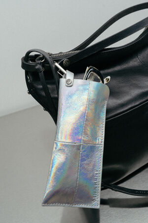 A pair of sunglasses in a shiny, iridescent case is attached to the handle of an INA KENT black leather handbag.
